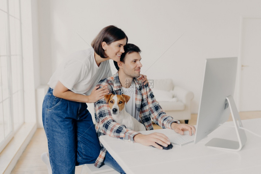 穿着白色t恤和牛仔裤的幸福妻子拥抱在计算机工作的丈夫帮助他编写报告重点是监视家用小狗在主人膝盖上做成的家用小狗图片