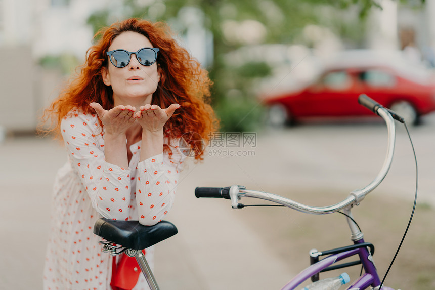 深爱的红发妇女向街上情人发出空吻将棕榈贴在嘴边穿圆窗帘衣服骑自行车在户外积极休息在街上摆姿势娱乐时间概念图片