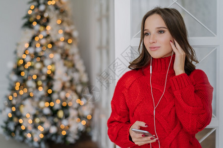 漂亮女利用手机和耳监听音乐站在家中反对圣诞节树灯背景并抄写文字空间图片