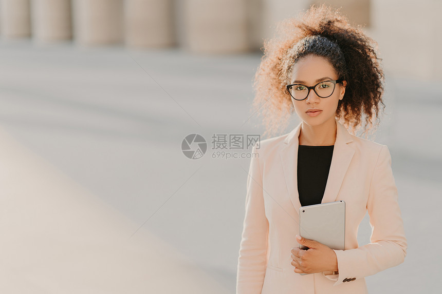 美籍高雅女商人使用平板电脑户外穿着正式的白色夹克透明戴黑色框的眼镜卷毛梳成马尾图片