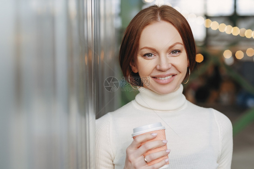 穿着白领毛衣的漂亮年轻女拿着纸杯的香味卡布西诺或咖啡笑得很开心的看着摄像机在外面摆姿势闲暇时间的概念图片