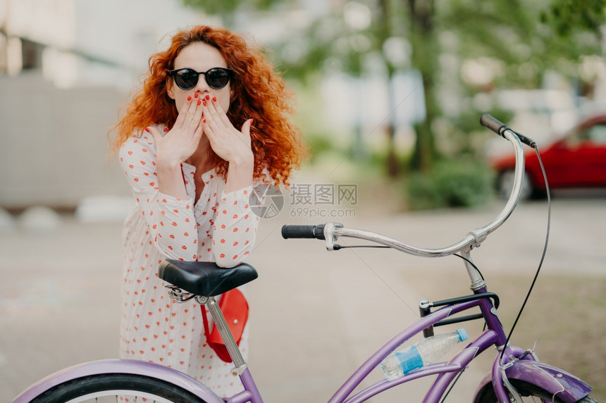 在户外拍摄华丽的红发女双掌盖着嘴部穿红色美甲着时装服戴和墨镜在自行车附近摆布免费骑自行车图片