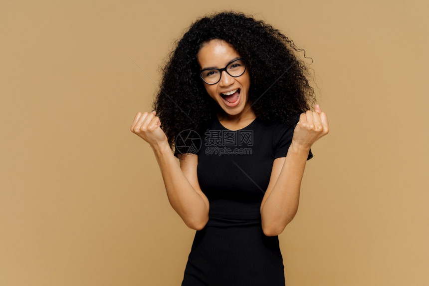 赢得胜利的可爱黑皮肤女人举起紧握的拳头欢乐地喊叫庆祝胜利穿黑色衣服站在棕背景上感觉自己是胜利者身体语言成功的概念图片