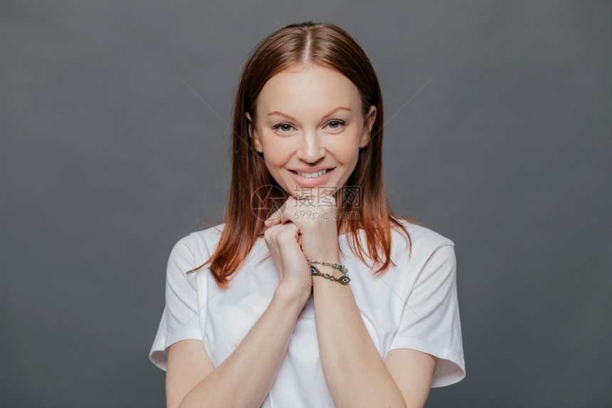 快乐的美丽女人横向镜头穿着随意的白色T衬衫保持双手在下巴微笑轻心情良好面对黑暗灰色工作室的背景正面情绪图片