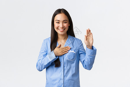 身穿蓝睡衣的诚实真和的亚洲女孩举起一只手将握在心上如许下诺言说出真相或发誓微笑如在白色背景上戴着果酱身穿蓝色睡衣举起一只手将握在背景