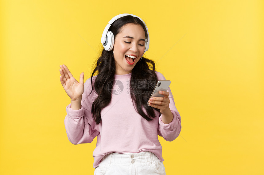 欢笑快乐和无忧虑的亚裔女孩在耳机上唱卡拉ok阅读手机的歌曲站立黄色背景人们的情感生活方式休闲和美丽概念欢笑快乐和无忧的亚裔女孩在图片