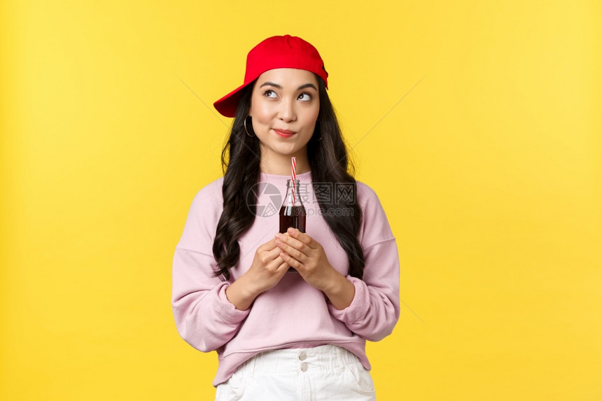 人们的情绪饮料和夏季休闲概念富有创意的时尚亚洲女孩穿着红色帽子笑像看左上角喝苏打水享受冷饮喝黄色背景富有创意的亚洲女孩穿着红色帽图片