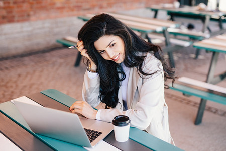 坐在户外使用笔记本电脑工作喝着咖啡试图制造项目可爱的黑发女工作图片