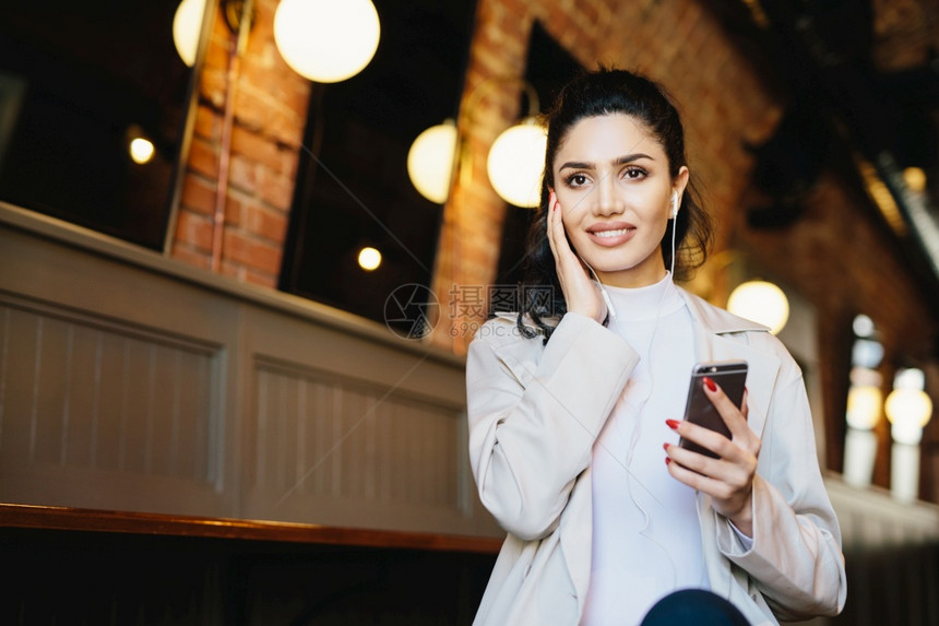 坐在咖啡厅里使用免费的互联网连接听用耳机和手的音乐使用现代技术的优雅女士图片