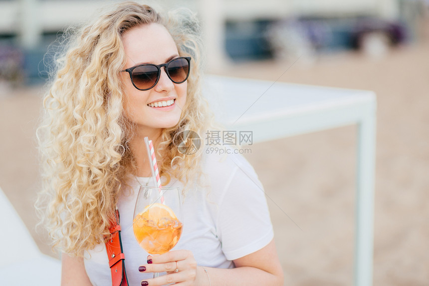 有卷发的正金色女郎戴太阳眼镜外表快乐微笑温柔鸡尾酒新鲜等海滩上的朋友图片