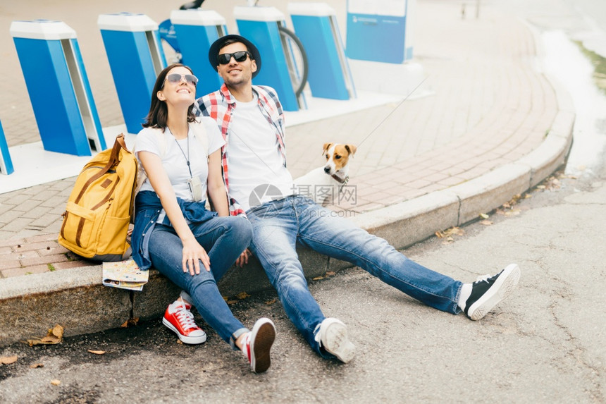 舒适的年轻男女旅行者及其宠物坐在人行道上享受良好天气的同时有愉快表情在公园长途跋涉之后疲惫不堪图片