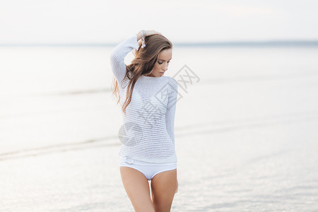 想着梦想进发穿着白色毛衣和比基尼的可爱年轻女模特儿站在美丽的海景面前看着深思熟虑梦想着在海滩上快乐的事物背景