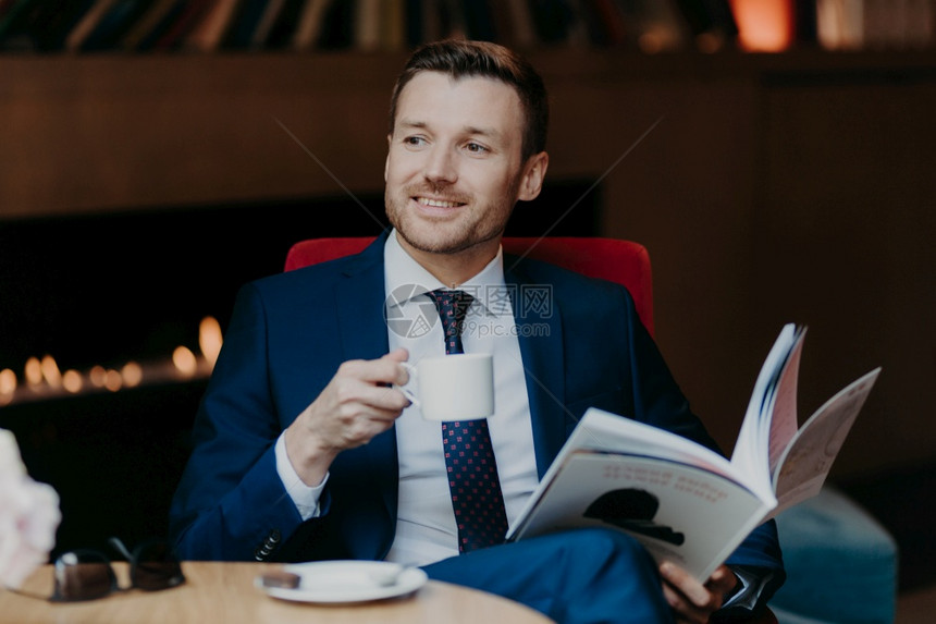 快乐的男经理或员工高兴的表达拿着咖啡躺在自助餐厅持有流行杂志专注意他的同事穿着正式衣服图片
