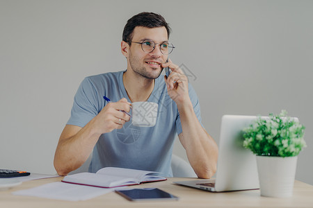 穿着圆杯眼镜的男会计师喜欢喝热饮拥有移动对话管理财务使用笔记本电脑在日中写笔图片