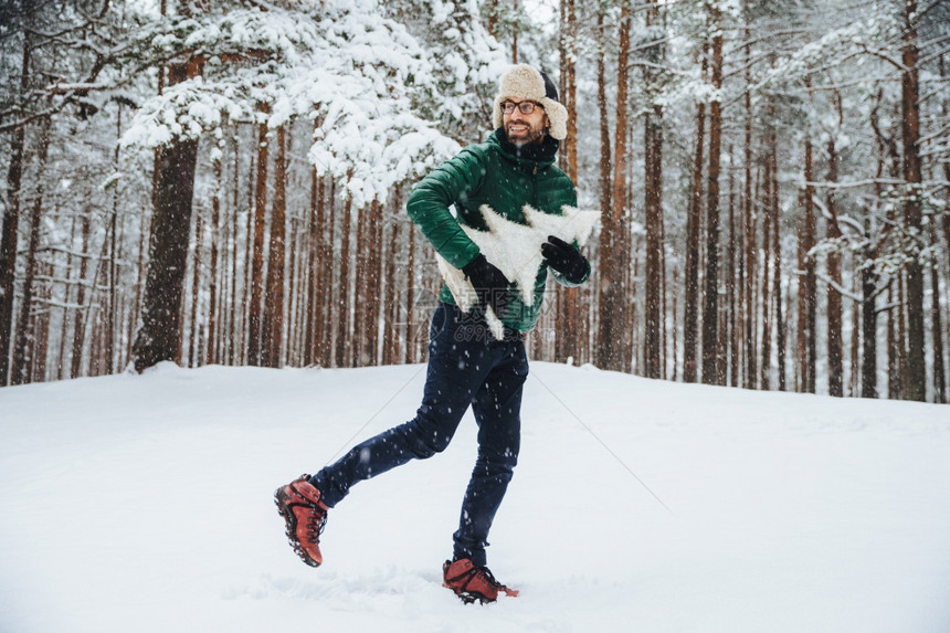 长着笑脸的英俊年轻男戴着温暖的帽子和动脉带在冬季准备节假日期间在森林外自由度假持有fir树图片