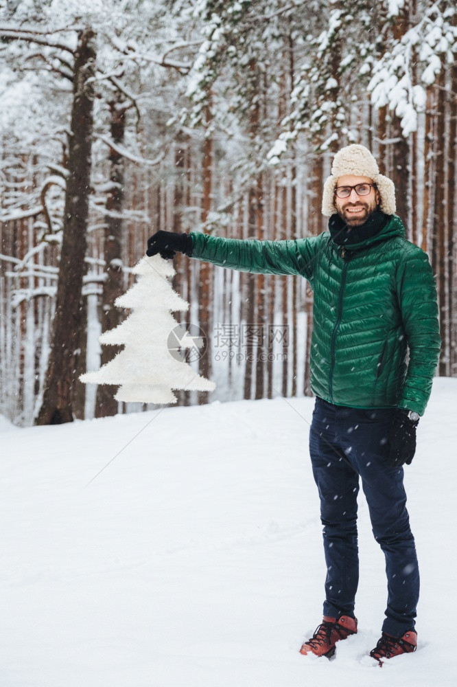 笑的男穿着时尚温暖衣服将白fir树握在手白色冬季森林中摆布图片
