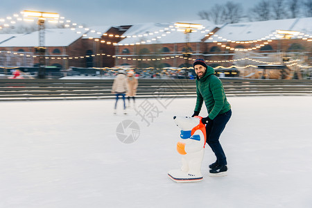 绿色夹克长胡子的男在户外玩新鲜空气享受寒冷的冬季天气在冰环上滑用Skae帮助害怕摔倒英俊的男人在滑冰环上玩得很开心背景
