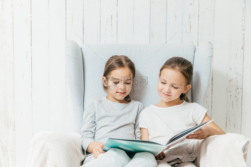 两位可爱的小姐妹坐在沙发上阅读有趣的书图片