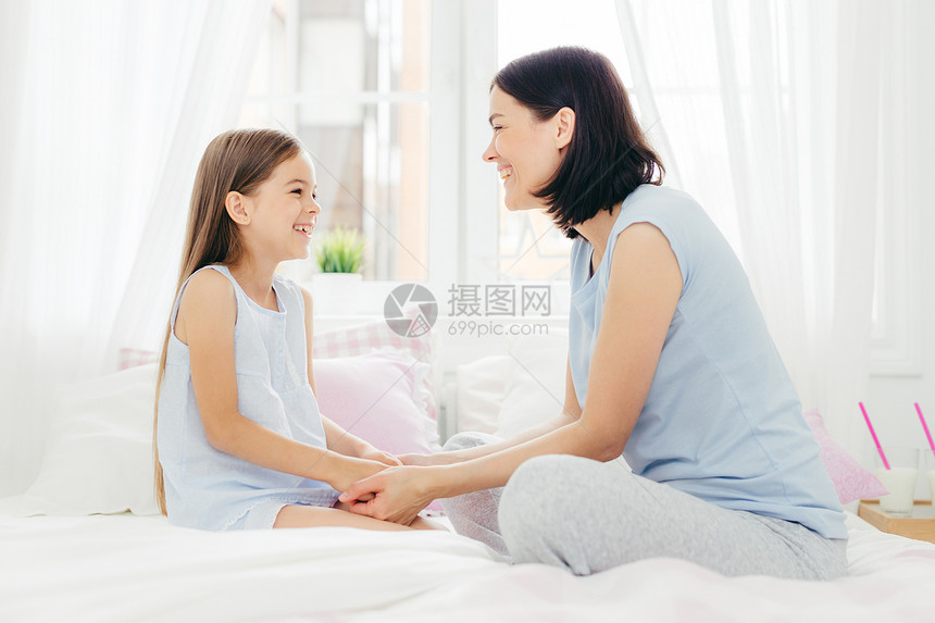 可爱的母亲和女儿享受在一起保持手愉快地聊天早上坐在舒适的床坐在轻的卧室保持良好的关系图片