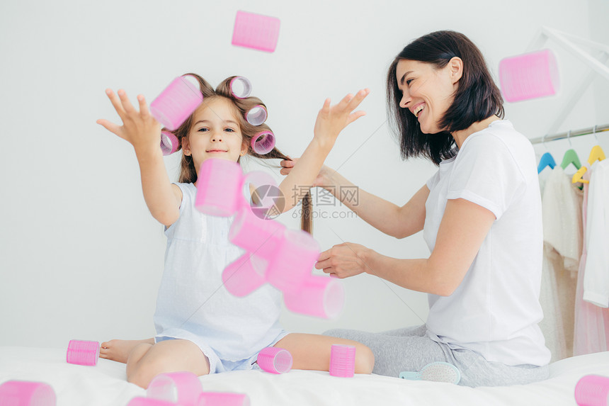 有趣的女孩在空气中扔卷发器一起玩乐她的母亲做发型穿便衣美丽的女人在头发上吹风坐在卧室里人家庭和母亲的概念图片