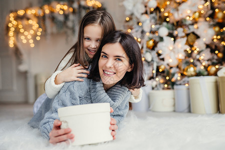 女孩和圣诞节这是给你的快乐小孩拥抱着她亲切的母带着包的礼物站在装饰背景与园林和新年树家庭气氛背景