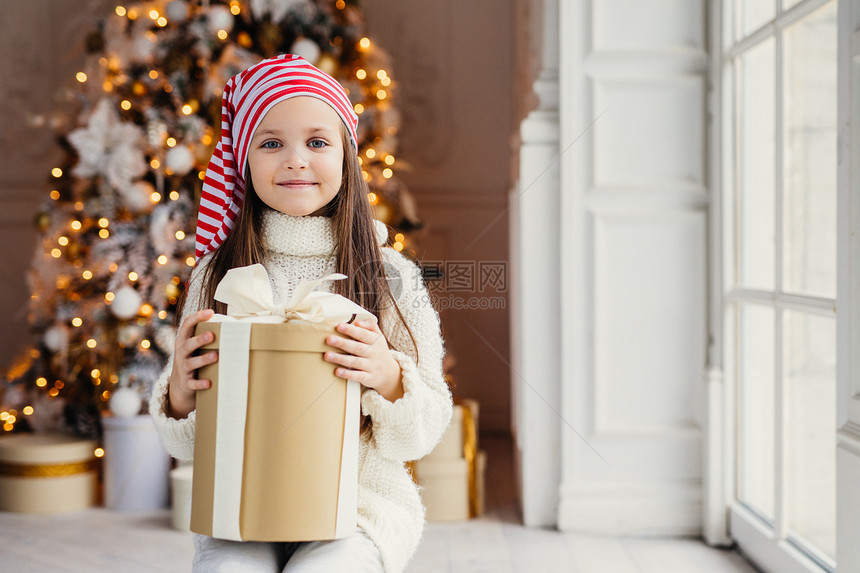 长着蓝色迷人的眼睛戴着圣诞帽子包装盒坐在饰的新年背景上坐过装饰的新年背景童庆祝概念图片