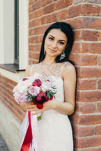 深色砖迷人的女新娘穿着白色婚纱拿花束站在砖墙附近有迷人的外貌背景