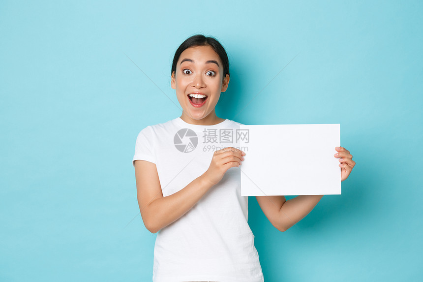 穿着白色t恤衫的亚洲女孩惊讶和兴奋做广告喘着惊讶展示标志一张空白纸与你的广告站着浅蓝色背景穿着白色t恤的亚洲女孩惊讶和兴奋站着浅图片