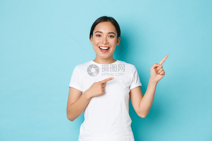 穿着白色t恤的美丽快乐亚洲女孩穿着白色t恤帮助选择展示道路或横幅韩国妇女满怀热情向右上角看蓝色背景美丽的亚洲女孩穿着白色t恤向右图片