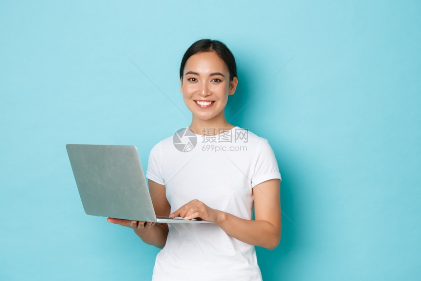 使用笔记本电脑在covid19自由或browingter期间在远程工作微笑着美丽的亚洲女孩在白色临时t恤短蓝背景上站立的肖像在c图片