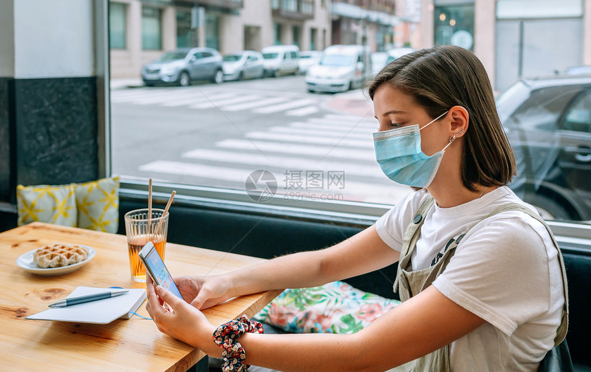 戴面罩的年轻妇女在咖啡店看手机时吃零食图片