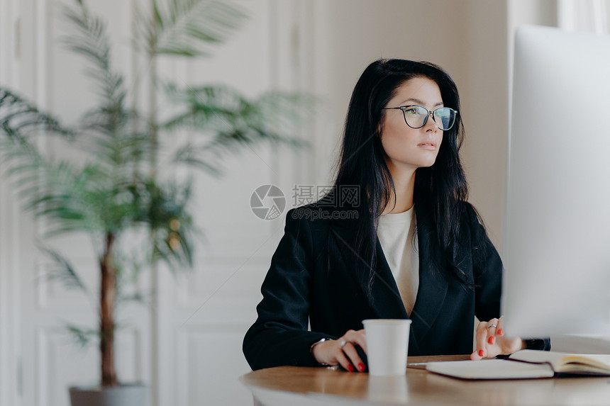 集中的女企业家穿着优雅的黑色服装在计算机工作坐在舒适场所的桌子上穿着透明的眼镜浏览互联网使商业项目考虑战略图片