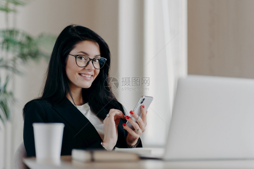 在现代智能手机上提供在线咨询微笑积极佩戴眼镜和正式西装坐在打开的笔记本电脑前图片