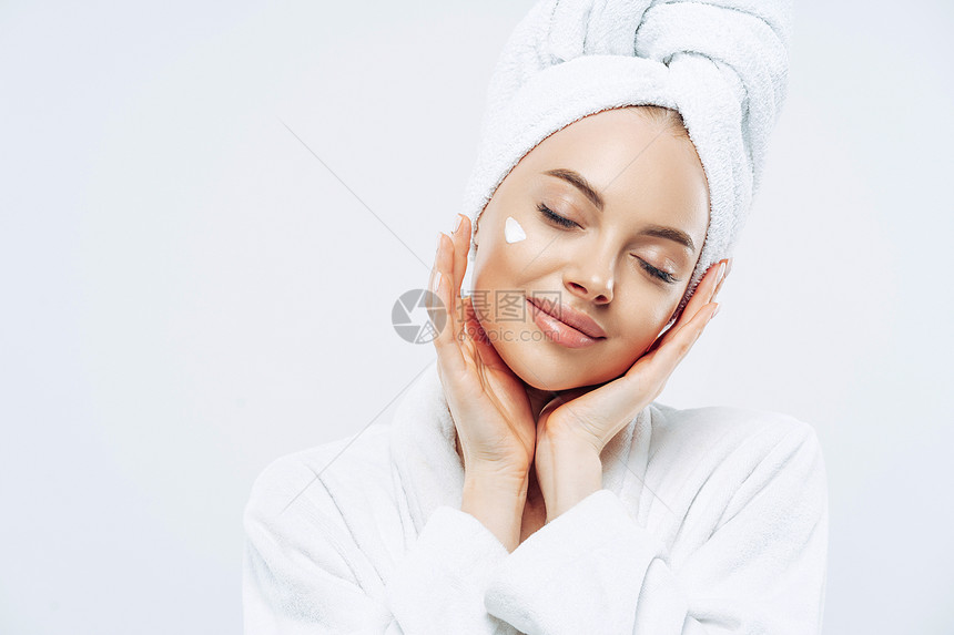 放松的美丽女士工作室的镜头闭着眼睛的美丽女士倾斜头部轻触皮肤施用面霜头部穿浴巾在做温泉疗程后享受面部治疗身体护理图片