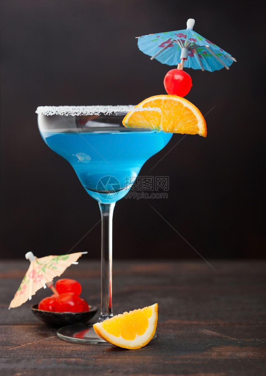 在玛格丽塔酒杯中装有蓝色环礁夏季鸡尾酒配有甜美鸡尾酒樱桃和橙色切片底桌有雨伞图片