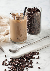 冰冷焦糖咖啡牛奶和玻璃容器豆子和勺在马贝尔板上和浅色桌底图片