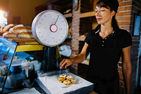 身穿黑色制服的美丽笑着容的女面包店助理以重量级甜烘烤食品为标准图片