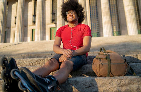 年轻拉丁男子用耳机听音乐和在户外溜冰后休息的肖像城市概念图片