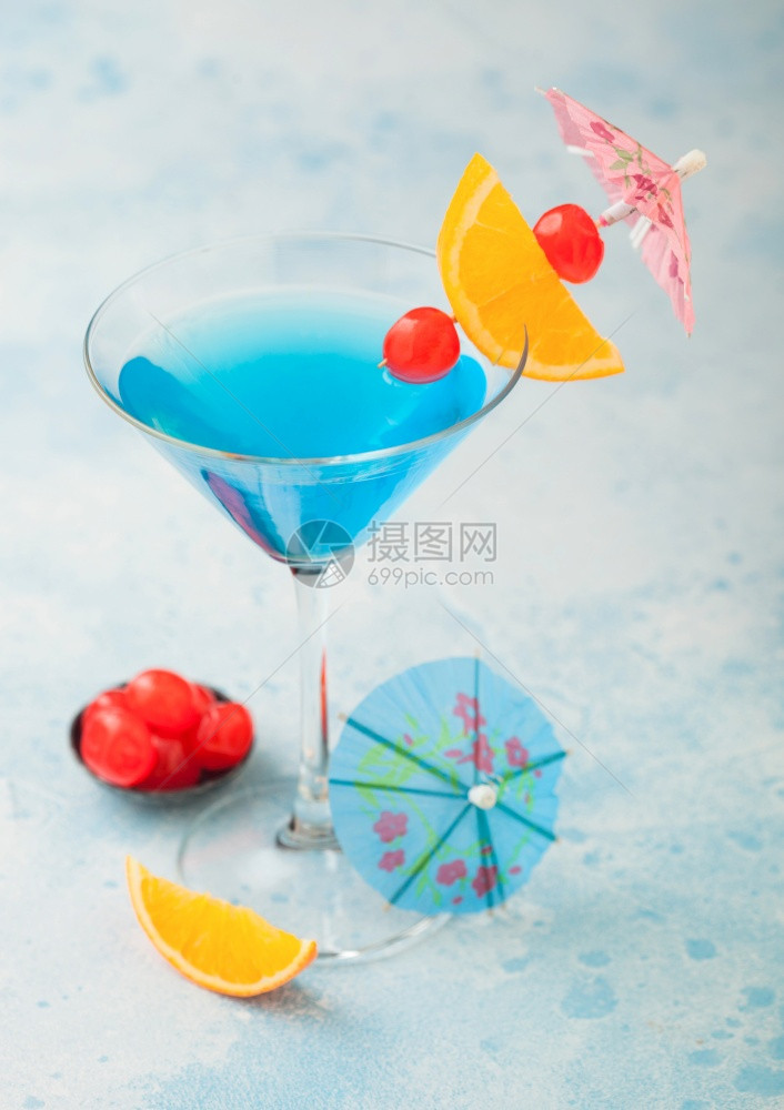 马提尼酒杯中的蓝色环礁夏季鸡尾酒配有甜樱桃和橙色切片蓝桌底有雨伞图片