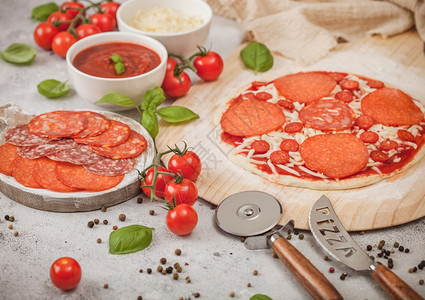 披萨刀准备烤番茄披萨配有沙拉米辣焦索配有车轮切割机新鲜西红柿和浅桌上的烤肉配有奶酪和番茄糊的碗盘背景