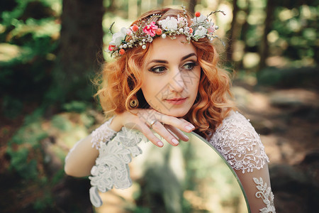 美丽的红发新娘在森林中头部和镜子上挂着花环夏天结婚日图片