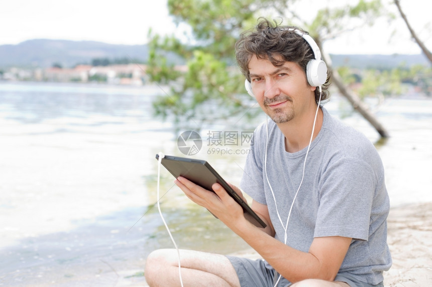 青年男子在海滩上拿着带耳机的平板电脑图片