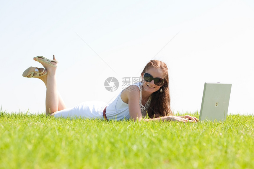 一个可爱的年轻女躺在公园的草地上用笔记本电脑图片