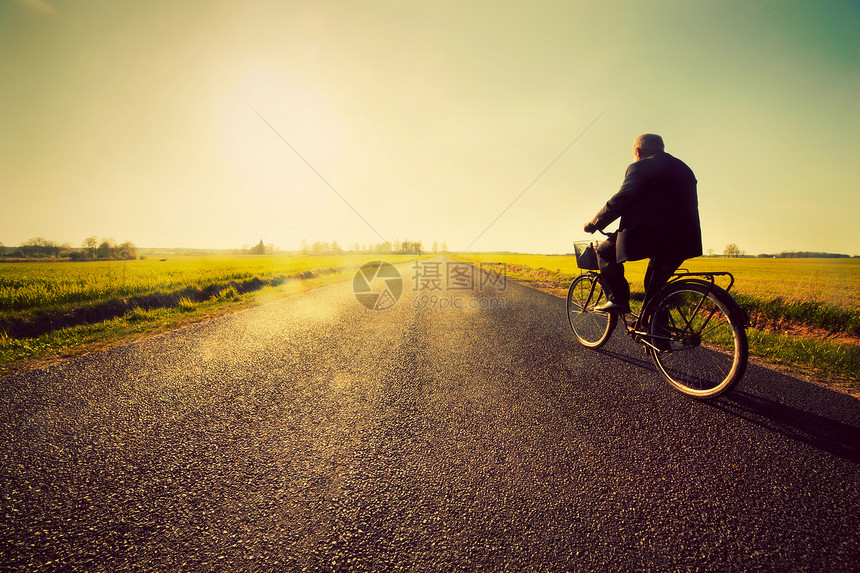 老人骑自行车在沥青路上骑自行车走向阳光的日落天空图片