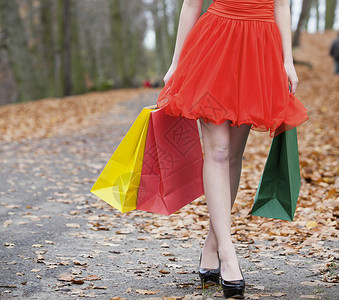 穿着优雅红色礼服的女孩穿着彩色的纸袋在秋天公园散步图片