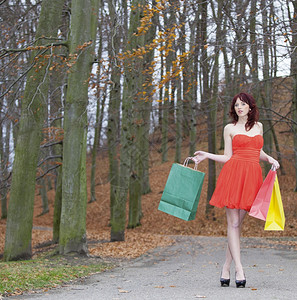 穿着优雅红色礼服的女孩穿着彩色纸袋在秋天公园散步图片