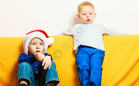 快乐节假日戴着红色圣塔帮手帽子的小男孩坐在沙发上图片