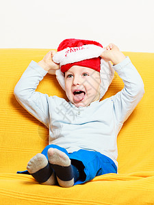 童年快乐节假日戴着红圣塔帮手帽子的小男孩坐在家里沙发上图片