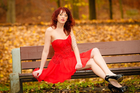 穿着优雅的红色礼服年轻美女坐在长椅上孩秋天公园放松背景图片