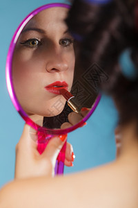 年轻女子准备参加聚会出去玩女孩用卷头发用红口涂看着镜子反光风格的蓝色背景背景图片